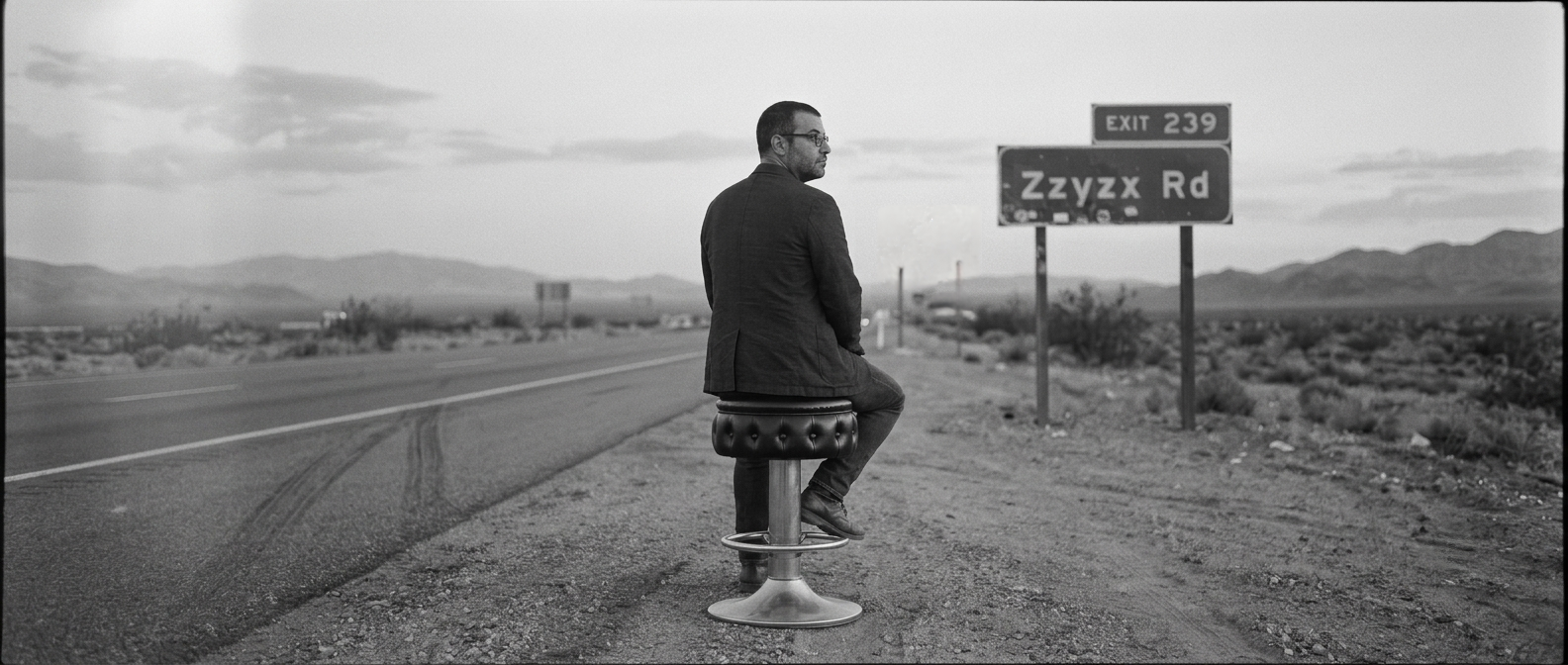 A man seated on a barstool along an empty desert highway, facing away from the camera, toward a road sign reading Exit 239, Zzyzx Rd.